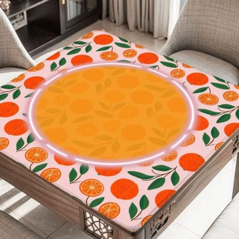 May include: A square table with a pink tablecloth featuring a pattern of oranges, orange slices, and green leaves. The center of the table has a large orange circle with a white glowing outline. The table is made of wood.