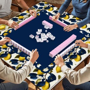 May include: A group of people playing Mahjong on a table. The table has a navy blue center and a decorative border with a lemon and leaf pattern. Pink tiles and dice are visible.