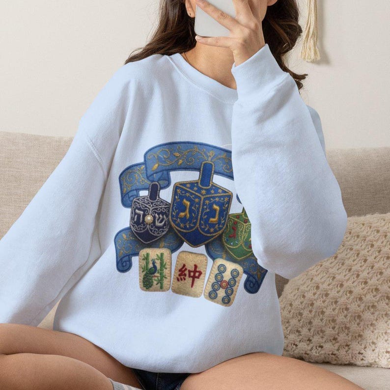 May include: A light blue crewneck sweatshirt featuring a graphic print of three dreidels centered on a blue ribbon banner, positioned above three mahjong tiles. The center dreidel displays a Hebrew letter, while the tiles show a peacock, a Chinese character, and a circular pattern.