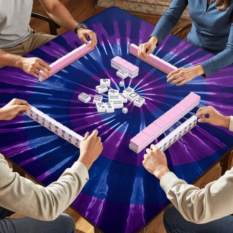 May include: A square mahjong table cover featuring a vibrant purple and blue tie-dye pattern. Four players are seated around the table, each with a rack of mahjong tiles. A collection of white mahjong tiles and two small dice are in the center.