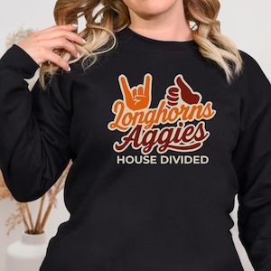 May include: Black sweatshirt with the text "Longhorns Aggies HOUSE DIVIDED" in orange and brown. The design includes hand gestures. A casual top for fans of college sports.
