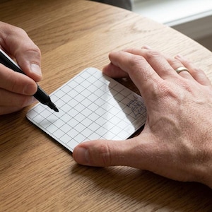 Peut inclure: Un sous-verre carré blanc avec un motif de grille noire et le texte "PEN AND LENS" imprimé dans le coin. Le sous-verre a une finition brillante et est utilisé avec un marqueur noir pour dessiner sur la surface de la grille.