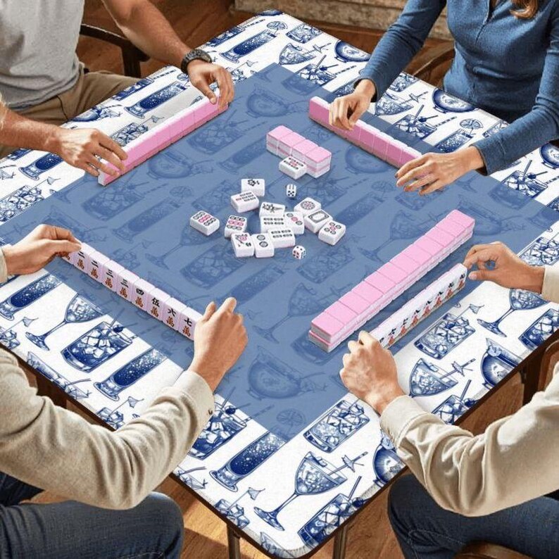 May include: A group of people playing mahjong on a blue table with a cocktail-themed border. The mahjong tiles are white with black markings, and the racks are pink. The table is covered with a blue cloth with a cocktail pattern.