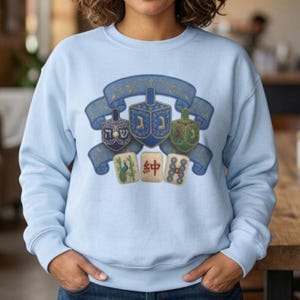 May include: A light blue crewneck sweatshirt featuring a graphic print of three dreidels centered on a blue ribbon banner, with three mahjong tiles positioned below the banner. The central dreidel displays Hebrew letters, while the tiles show a peacock, a red character, and circles.