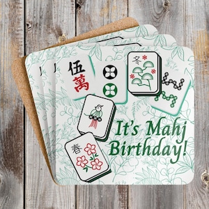 May include: A stack of square coasters with a cork backing. The coasters feature a white background with green floral patterns and various Mahjong tile designs. The text "It's Mahj Birthday!" is printed in green.