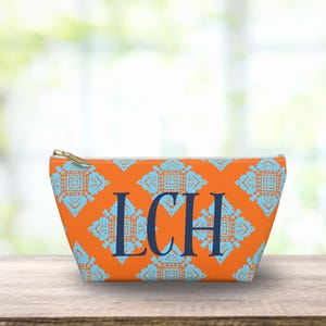 May include: Orange cosmetic bag with a repeating turquoise geometric pattern. The bag features the initials "LCH" in navy blue. It has a gold zipper and is sitting on a wooden surface.
