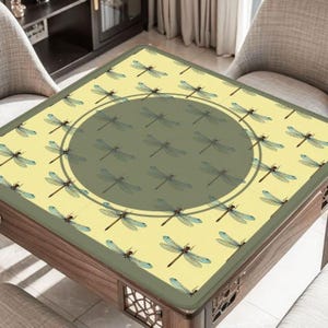 May include: A square table with a light yellow surface featuring a dragonfly pattern. The center has a large, dark green circle with more dragonflies. The table has a dark wood frame and decorative cutouts.