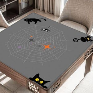 May include: A gray Halloween-themed table cover featuring a spider web design with a spider, black cats, and starburst accents. The table cover is rectangular and sits on a wooden table.