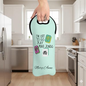 May include: A light teal wine bottle tote bag with a black handle. The bag features the text "I'M OFF TO GO PLAY MAH JONGG" along with colorful Mahjong tiles and the name "Mary Anne".
