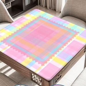 May include: A square table with a pastel-colored, plaid-patterned tablecloth. The tablecloth features a pink center, surrounded by layers of blue, yellow, and orange squares. The table has a dark wood frame and is set in a room with neutral-toned chairs.