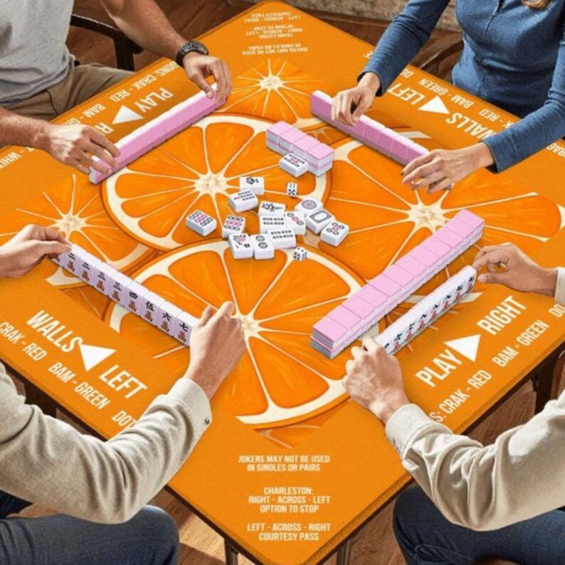 K&ouml;nnte beinhalten: Ein quadratischer, orangefarbener Mahjong-Tisch mit gro&szlig;en Orangenscheiben-Designs. Die Spieler halten rosa und wei&szlig;e Spielsteine. Der Tisch tr&auml;gt die Aufschrift "Walls Left" und "Play Right".