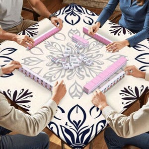 May include: A square game table with a floral design is surrounded by people playing Mahjong. The table has a cream-colored surface with a large gray and navy blue floral pattern. Pink Mahjong tiles are arranged around the table.