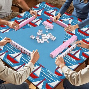 May include: A group of people playing Mahjong on a blue table. The table has a blue surface in the center and a patterned border with red sailboats on a white and blue background. Pink Mahjong tiles and racks are visible.