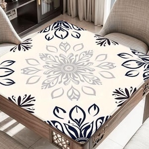 May include: A square wooden table with a cream-colored tablecloth featuring a large, symmetrical floral design in shades of gray and navy blue. The table has a dark wood frame and is surrounded by two beige chairs.