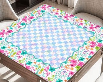 May include: A square wooden table with a floral patterned top. The top features a light blue and white checkered center, framed by a scalloped edge and a border of colorful flowers in pink, green, and orange.