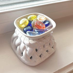 May include: A white ceramic oil warmer with a woven design base, topped with a small dish filled with colorful glass stones. The stones are yellow, blue, orange, and clear.