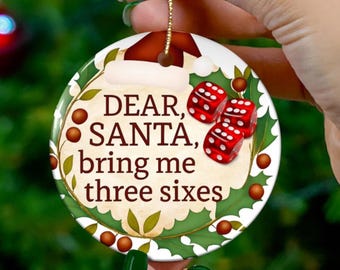 May include: Round Christmas ornament with the text "DEAR, SANTA, bring me three sixes." The ornament features red dice, holly, and a Santa hat design. A gold string is attached for hanging.