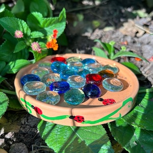 May include: A round terracotta saucer featuring a hand-painted green vine motif with small red ladybugs. The interior of the saucer is filled with a collection of smooth, translucent glass gems in various shades of blue, red, and clear glass.