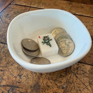 May include: A white, oval-shaped bowl containing several coins and a white Mahjong tile with the number 1 and a green and red design. The bowl sits on a wooden surface.