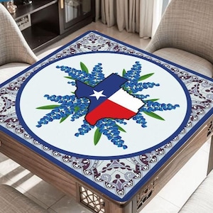 May include: A square table cover featuring a Texas state outline in blue, white, and red, surrounded by bluebonnet flowers. The design is centered on a white background, framed by a blue border with a decorative pattern. The table is made of wood.