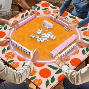 May include: A Mahjong game in progress, featuring a table with an orange and green citrus fruit pattern. The game set includes pink tile racks and white tiles with black symbols. Players are seen interacting with the game pieces.