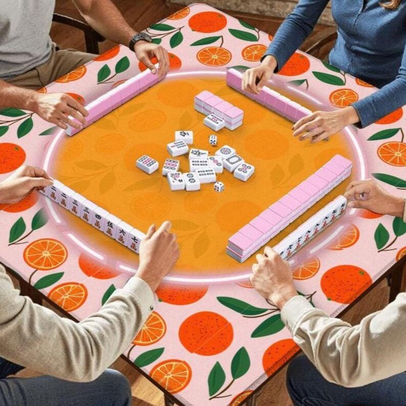 May include: A Mahjong game in progress, featuring a table with an orange and green citrus fruit pattern. The game set includes pink tile racks and white tiles with black symbols. Players are seen interacting with the game pieces.