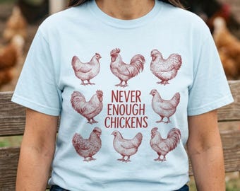 May include: A light blue short-sleeve t-shirt featuring a graphic print of eight different chickens arranged in a circle around the dark red text that reads NEVER ENOUGH CHICKENS. The design is printed in a dark red ink on the front of the shirt.