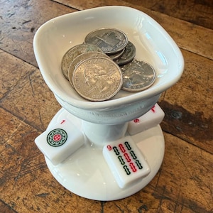 May include: A white ceramic coin holder with a bowl-shaped top, holding several silver coins. The holder is supported by a pedestal base with three attached Mahjong tiles. The tiles are white with red and green markings.