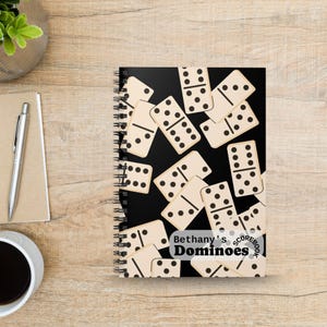 May include: A spiral-bound notebook with a black cover featuring a scattered pattern of cream-colored dominoes with black pips. A white rectangular label on the bottom right corner displays the text "Bethany's Dominoes SCOREBOOK" in black.