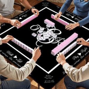 May include: A black square mahjong table mat featuring a white border and central decorative scrollwork. The mat includes printed text labels for game directions and positions. Pink mahjong tiles are arranged in rows along the edges.