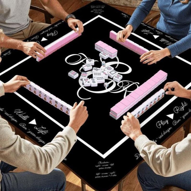 May include: A black square mahjong table mat featuring a white border and central decorative scrollwork. The mat includes printed text labels for game directions and positions. Pink mahjong tiles are arranged in rows along the edges.
