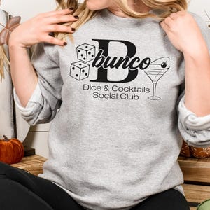 May include: A light gray sweatshirt with the word "bunco" in a black script font, along with dice and a cocktail glass graphic. The text "Dice & Cocktails Social Club" is below the graphic.