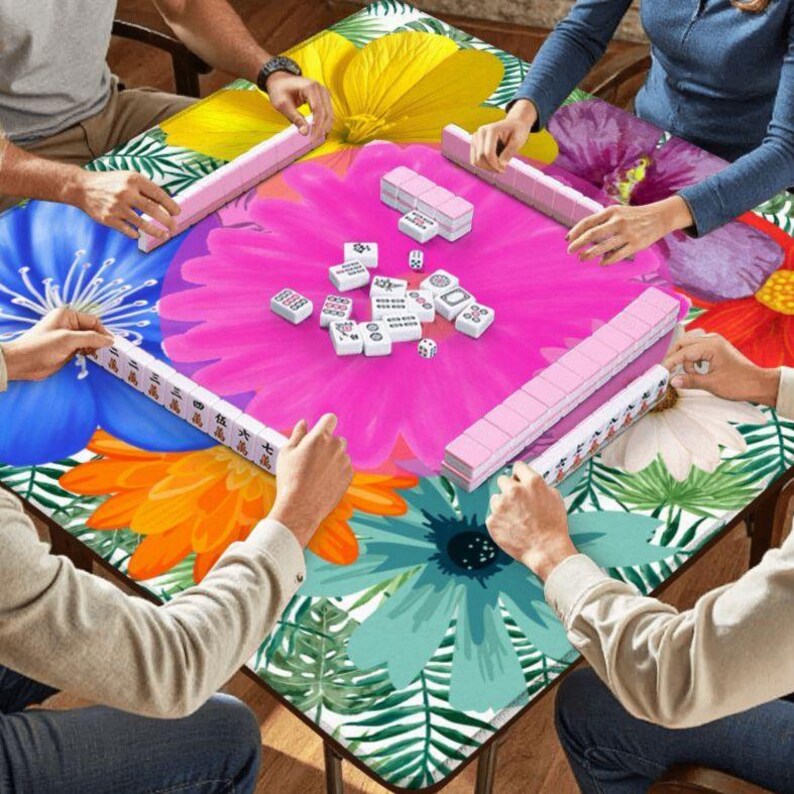 May include: A square Mahjong table with a floral design featuring large flowers in yellow, blue, orange, and pink. Players are seated around the table, holding pink Mahjong tiles and racks. White Mahjong tiles and dice are in the center.