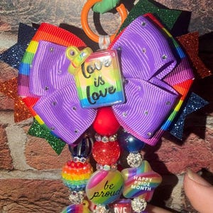 May include: A decorative keychain featuring a large purple bow with rainbow-striped edges and glittery star points. Dangling from the bow are multiple strands of colorful beads, including star and heart shapes with text like Love is Love.
