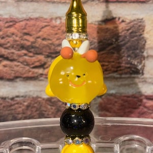Puede incluir: Un adorno para bolígrafo con forma de abeja amarilla con antenas naranjas y alas blancas, situado sobre una alternancia de cuentas negras brillantes y cuentas amarillas sólidas, separadas por espaciadores plateados con pedrería transparente.