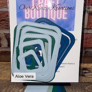 May include: A package of layered paper die cuts in shades of blue, green, and white. The die cuts are square with rounded corners. The package has the text "Craft Boutique" and "Our Krazy Kreations". The label says "Aloe Vera".