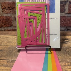 May include: A set of craft dies in a clear plastic package with a white background. The dies are in the shape of frames and are arranged in a stack. The package has the text "CRAFT Our Krazy Kreations" printed on it. The dies are pink and green. There are also six sheets of cardstock in different colors: pink, blue, green, orange, and red.