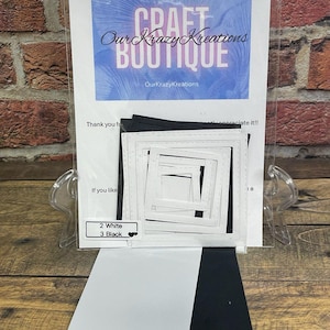 May include: A craft kit with white and black square die-cut frames. The kit includes a sheet with the text "Craft Boutique Our Krazy Kreations" and a label indicating 2 white and 3 black frames. The background is a brick wall.