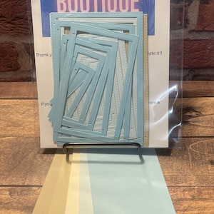 May include: A set of blue paper crafting dies in the shape of nested squares. The dies are packaged in a clear plastic bag with a white cardstock backing. The cardstock has the text "Our Krazy Kreations Craft Boutique" printed on it. The dies are displayed on a black stand with a set of beige and blue cardstock samples.