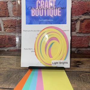 May include: A set of six die-cut paper circles in various shades of yellow, pink, blue, and green. The circles are arranged in a concentric pattern, with the largest circle at the bottom and the smallest circle at the top. The circles are labelled "Light Brights".