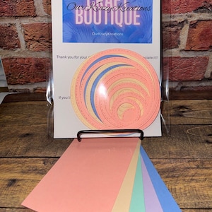 May include: A set of six paper crafting dies in various pastel colors, including peach, yellow, green, lavender, and blue. The dies are shaped like concentric circles and are packaged in a clear plastic bag with a white card that reads "CRAFT Our Krazy Kreations BOUTIQUE OurKrazyKreations Thank you for your appreciate it!! If you li"