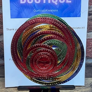 Puede incluir: Un conjunto de 10 troqueles de círculo metálico en rojo, verde, dorado y azul con un borde cosido. Los troqueles están dispuestos en un patrón circular. Los troqueles están empaquetados en una bolsa de plástico transparente con una tarjeta blanca que dice "Our Krazy Kreations Craft Boutique".