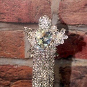 May include: A silver-toned decorative hair accessory featuring a central heart-shaped crystal, a rhinestone-encrusted head, and wings. The piece is adorned with small white faux pearls and a flower accent, with multiple long, dangling rhinestone fringe strands.