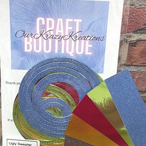 Puede incluir: Un kit de manualidades con recortes de papel circulares azules, verdes y rojos en capas, junto con una selección de cartulinas brillantes y metálicas de colores. El kit incluye un clip y una tarjeta con el texto "Craft Boutique" y "Ugly Sweater".