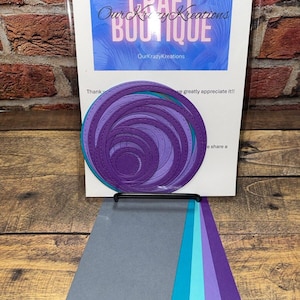 May include: A set of layered paper die cuts in shades of purple and teal, arranged in a spiral pattern. The set includes a variety of colored cardstock samples. The background features a sign with the text "CRAFT BOUTIQUE" and "OurKrazyKreations".