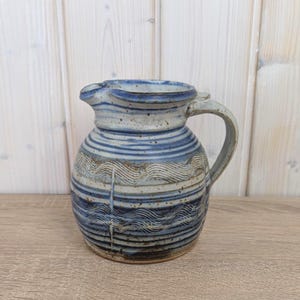 Könnte beinhalten: Handgefertigter Keramikkrug mit blau-weißem Streifendesign. Der Krug hat einen Henkel und einen Ausguss zum Ausgießen. Das Design umfasst wellenförmige Linien und horizontale Streifen. Ein dekorativer Artikel für Küche oder Esszimmer.