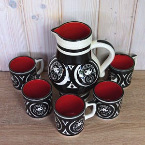 May include: A black and white ceramic pitcher and matching mugs set. The pitcher and mugs have a red interior. The set features a repeating design of stylized faces and geometric shapes. The pitcher has a handle and spout.
