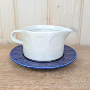 May include: A white ceramic creamer with a handle and a spout, decorated with embossed circles. It rests on a deep blue saucer, also with a circle design. Ideal for serving beverages, this set is a classic design.