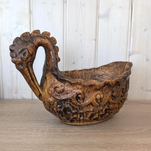 May include: A brown ceramic planter with a bird-head handle. The planter features a textured surface with decorative swirls and patterns. The color is a mix of brown and black, with a glossy finish. It is designed for plants or flowers, a decorative item.
