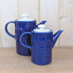 May include: Two cobalt blue ceramic teapots with white lids. The teapots have a repeating circle pattern and curved handles. The teapots are of different sizes and are displayed on a wooden surface.